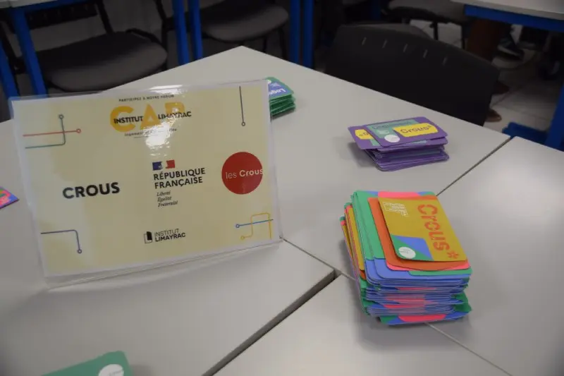 Présentoir de table arborant le logo du Crous à côté de piles de cartes d'information colorées disposées sur un bureau.
