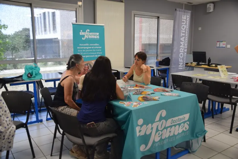 ne conseillère assise à une table couverte d'une nappe bleue Info Jeunes échange avec deux personnes venues s'informer lors d'un forum.