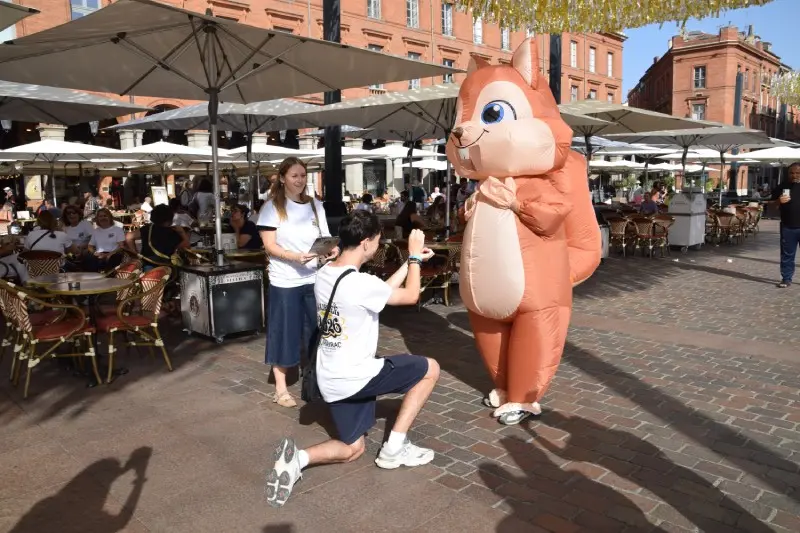 Un étudiant à genoux fait une demande factice à la mascotte écureuil géante sur la place du Capitole lors d'un événement festif.