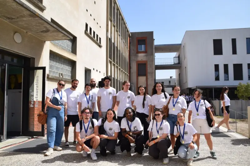 Groupe de jeunes étudiants souriants portant des t-shirts blancs identiques et des médailles autour du cou devant un bâtiment scolaire.